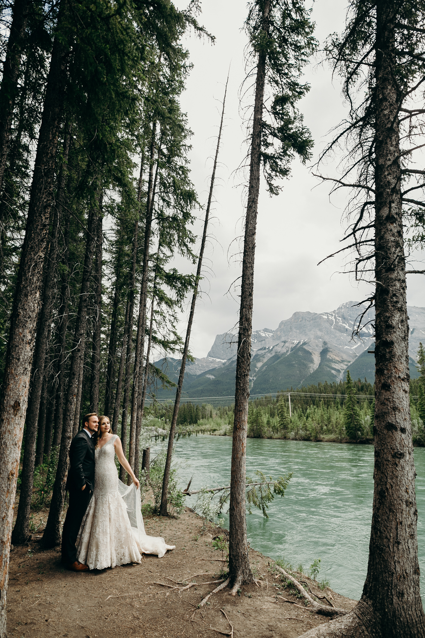 Bride and groom stand on cliff with river in background and fallen tree Canmore Bow River