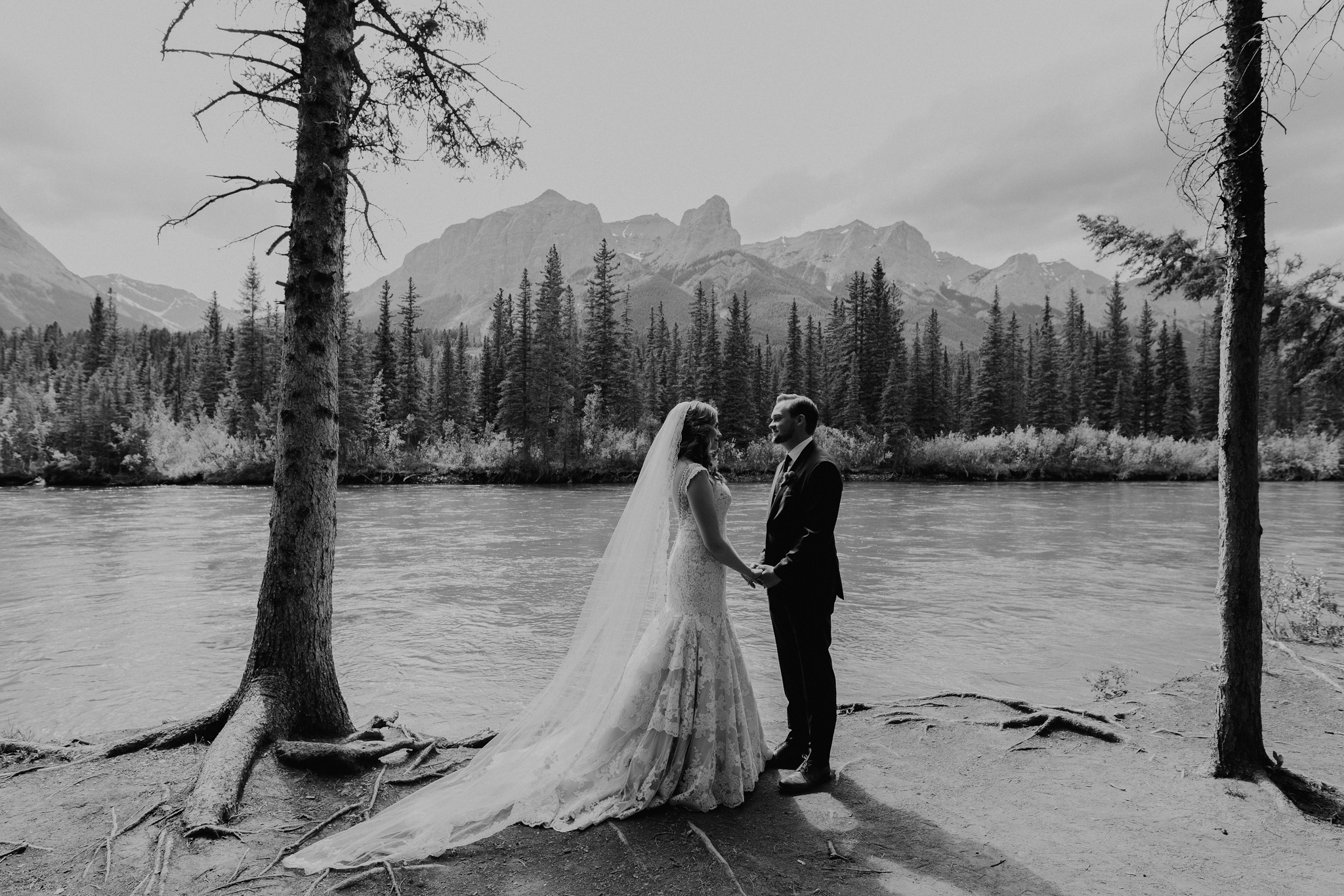 Bride and groom stand on cliff with river in background and fallen tree Canmore Bow River