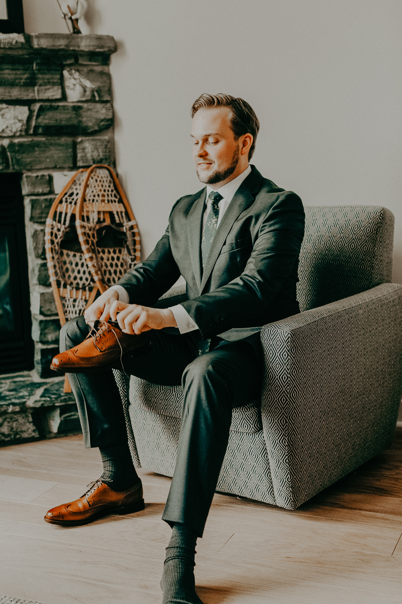 Groom sits on grey chair in grey suit and puts on brown dress shoes