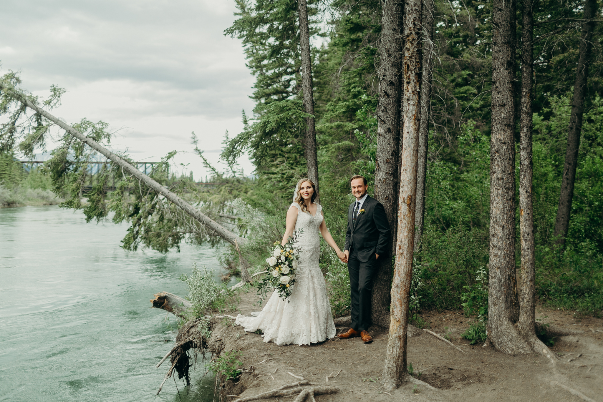 Bride and groom stand on cliff with river in background and fallen tree Canmore Bow River